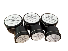 Load image into Gallery viewer, Six small black leather blocks with round white labels reading Dungeon, arranged on a plain white background. The top labels show different scent notes: Amber Vanilla, Bourbon Vanilla and Leather.